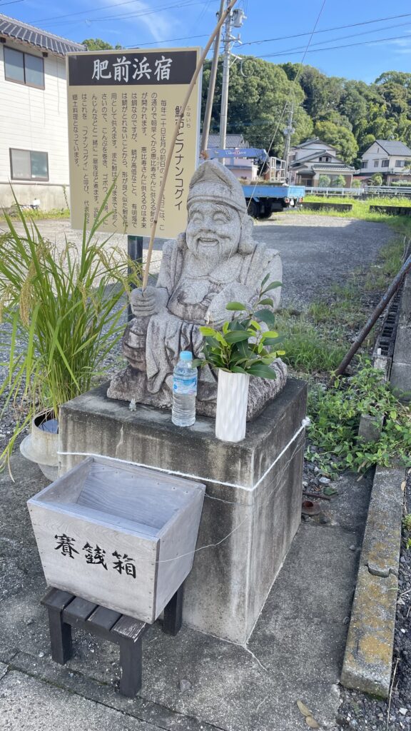Stone statue of Ebisu deity with a fishing rod at Hizen-Hamashuku in Kashima, Saga, with offering box and plants beside it.