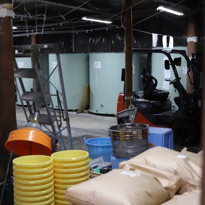 Brewing Floor at Mitsutake Sake Brewery Brewing work area at Mitsutake Sake Brewery in Kashima, Saga—stacks of rice sacks, yellow buckets, stainless tools, and large fermentation tanks with a forklift in the background.